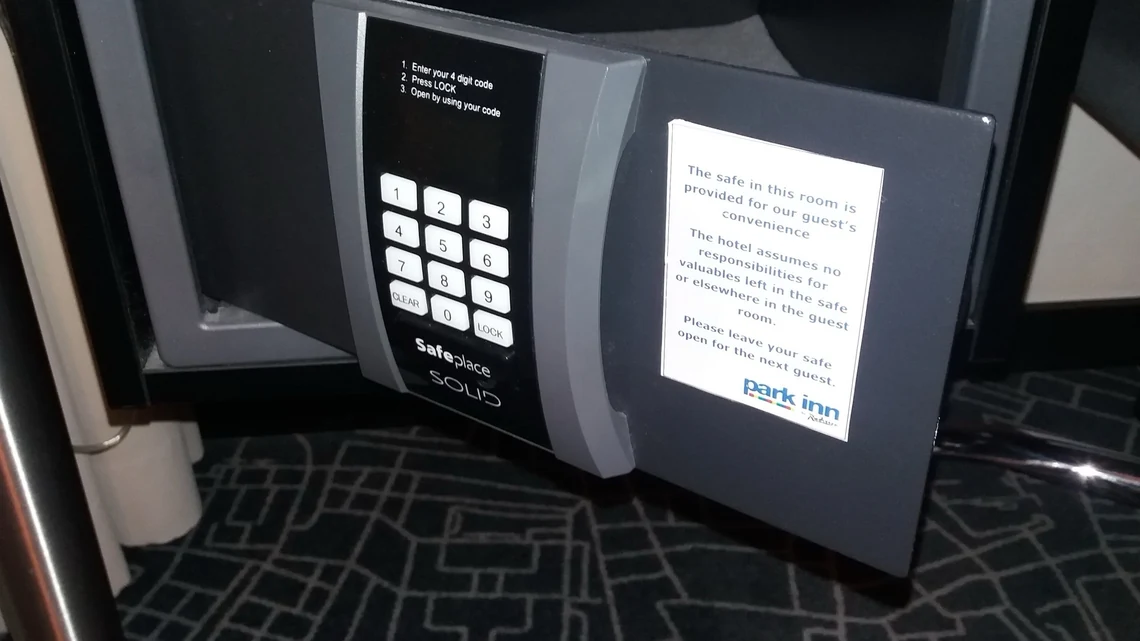Bir otel odasındaki küçük çelik kasa açık duruyor; kapağın iç yüzünde kullanım talimatı ve tuş takımı görünüyor.