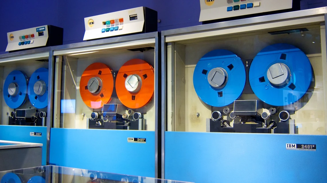 Bir müzede cam vitrin ardında sıralanmış IBM 2401 manyetik teyp üniteleri; kırmızı ve mavi teyp makaraları takılı duruyor.